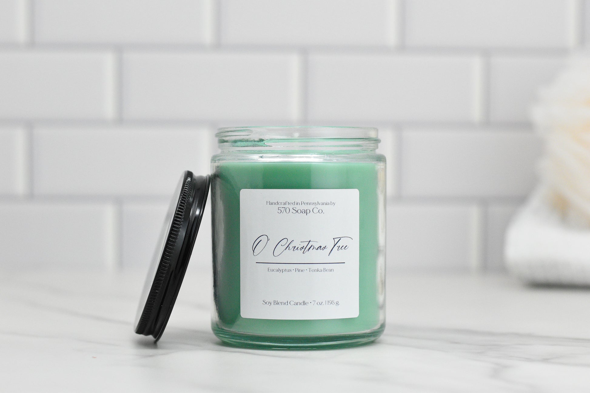 The 570 Soap Co. O Christmas Tree soy blend candle, in a clear jar with a black lid, exudes a Winter Forest aroma of eucalyptus, pine, and tonka bean. It sits on white marble against subway tiles with a folded towel in view.