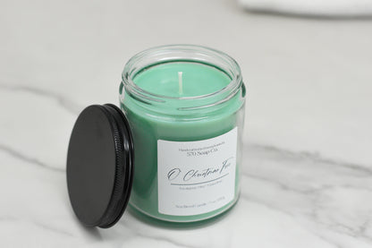 The 570 Soap Co. O Christmas Tree green soy blend candle is in a clear glass jar with a black lid, labeled with scent notes like Winter Forest Aroma, and displayed on a white marble surface.