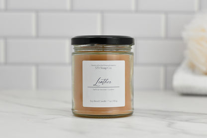 A glass jar candle called the Leather Soy Blend Candle from 570 Soap Co., featuring a black lid and crafted with toxin-free wax, emits a luxurious saffron and leather fragrance. It is beautifully displayed on a marble surface against a white tile backsplash.