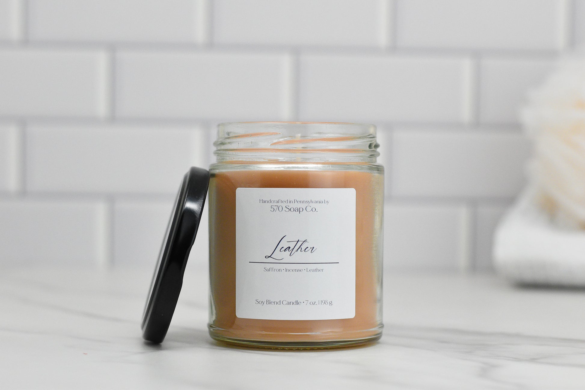 A glass jar candle labeled "Leather Soy Blend Candle" from 570 Soap Co. sits elegantly on a marble surface. The lid is slightly open, revealing a light brown toxin-free wax that emits a luxurious scent. In the background, a white tile wall and a blurred, soft object complete the soothing scene.