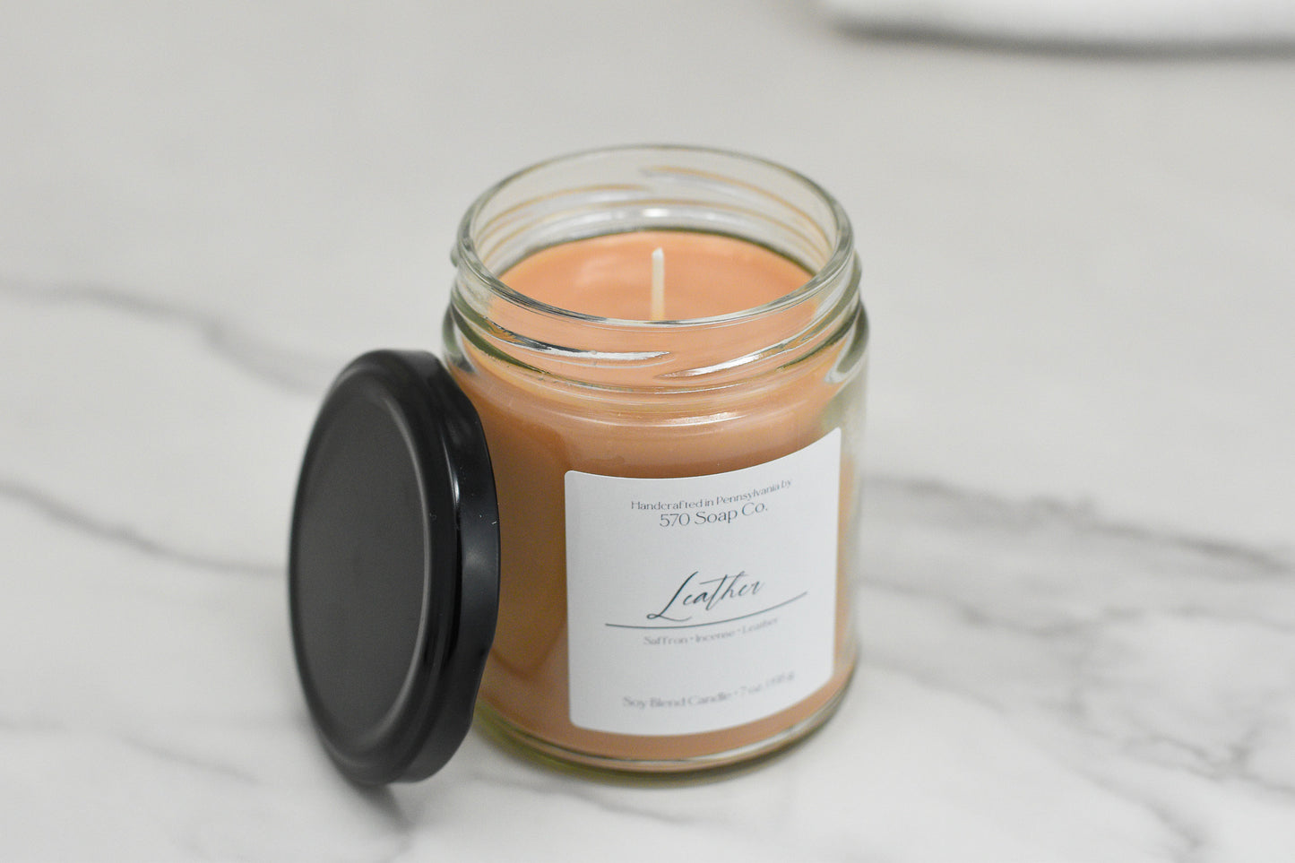 A glass jar holds a peach-colored Leather Soy Blend Candle, accompanied by a black lid. The label on the jar reads Leather, showcasing its creation by 570 Soap Co. Made from toxin-free wax, this luxurious scented candle is beautifully displayed on a marble surface.