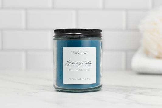 A blue candle in a glass jar with a black lid rests on a marble surface. The label displays 570 Soap Co., presenting their Blueberry Cobbler Soy Blend Candle with scent notes of Fresh-Baked Aroma, featuring blueberry, vanilla, and bakery dough. The setting is finished with a white tile background.