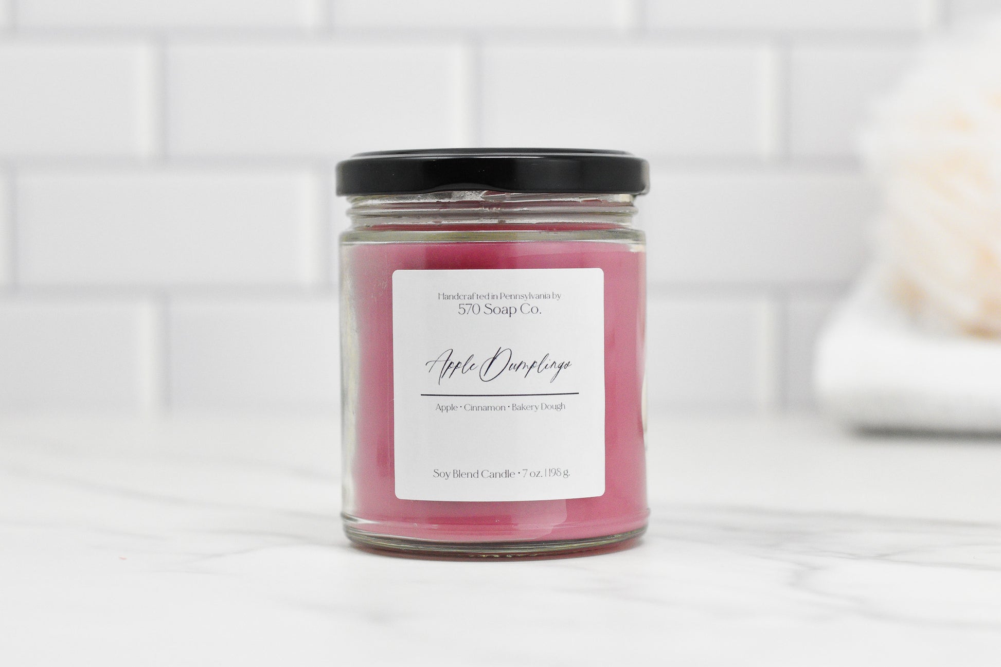 A pink Apple Dumpling Soy Blend Candle by 570 Soap Co. rests on white marble, encased in a clear jar with a black lid. Its elegant label reads Apple Cinnamon Bakery Dough. The toxin-free wax provides a clean burn as sweet apple notes fill the air against white subway tiles.