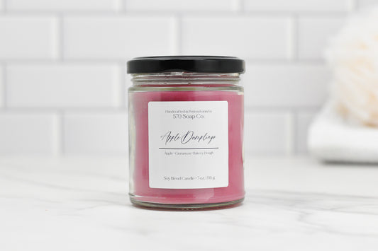 A pink Apple Dumpling Soy Blend Candle by 570 Soap Co. rests on white marble, encased in a clear jar with a black lid. Its elegant label reads Apple Cinnamon Bakery Dough. The toxin-free wax provides a clean burn as sweet apple notes fill the air against white subway tiles.