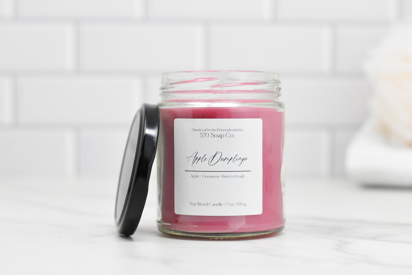 A pink Apple Dumpling Soy Blend Candle by 570 Soap Co. labeled Apple Overflows sits on marble. Made with toxin-free wax, it features a glass jar with a black lid beside it against white subway tiles.