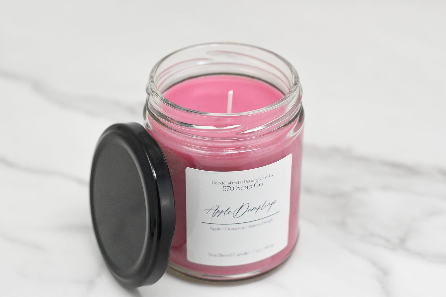 A pink Apple Dumpling Soy Blend Candle by 570 Soap Co. sits on a marble surface in a clear jar with a black lid. Labeled Apple Orchard, it highlights apple and cinnamon notes and uses toxin-free wax for a safe, cozy ambiance.