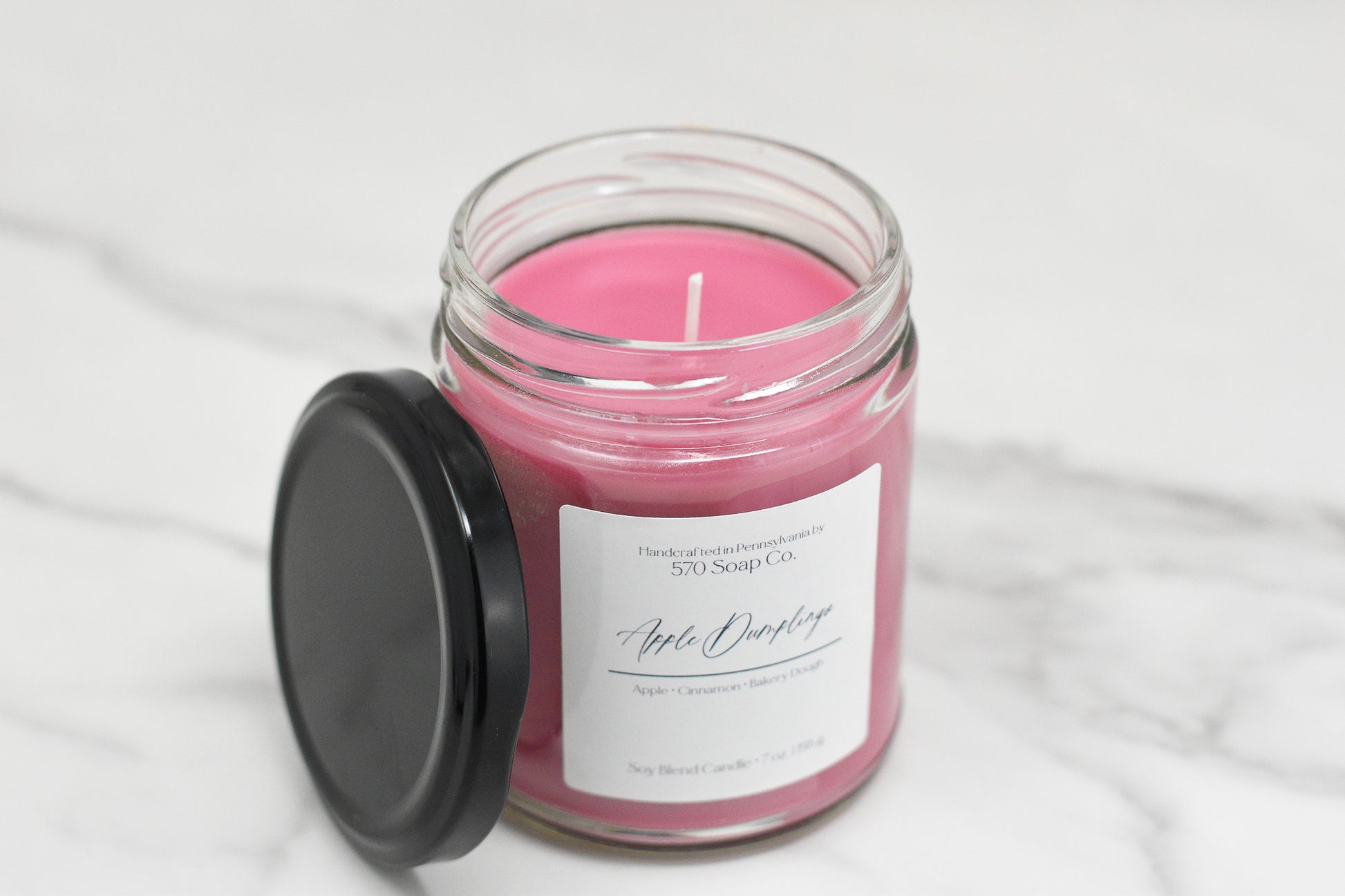 A pink Apple Dumpling Soy Blend Candle by 570 Soap Co. sits on a marble surface in a clear jar with a black lid. Labeled Apple Orchard, it highlights apple and cinnamon notes and uses toxin-free wax for a safe, cozy ambiance.