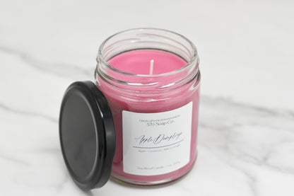 A pink Apple Dumpling Soy Blend Candle by 570 Soap Co. sits on a marble surface in a clear jar with a black lid. Labeled Apple Orchard, it highlights apple and cinnamon notes and uses toxin-free wax for a safe, cozy ambiance.