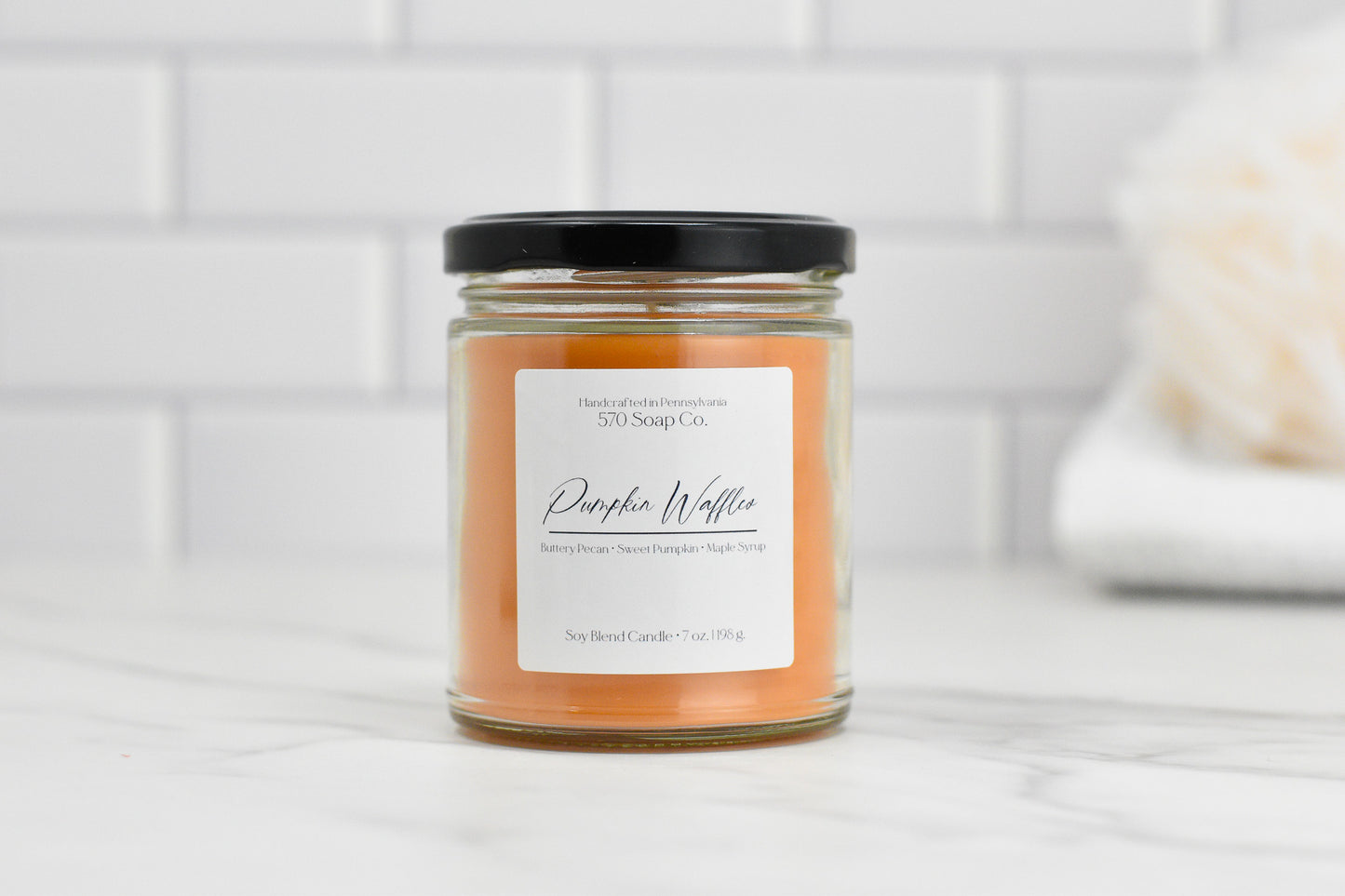 A small glass jar candle from 570 Soap Co., featuring amber-colored wax and a black lid, is labeled as the Pumpkin Waffles Soy Blend Candle. It emits a cozy aroma of butterscotch pecan, sweet pumpkin, and maple syrup. Placed against a white tiled background, this candle brings warmth to any space.