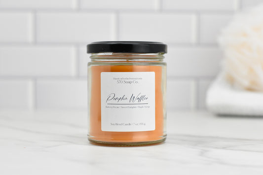 A small glass jar candle from 570 Soap Co., featuring amber-colored wax and a black lid, is labeled as the Pumpkin Waffles Soy Blend Candle. It emits a cozy aroma of butterscotch pecan, sweet pumpkin, and maple syrup. Placed against a white tiled background, this candle brings warmth to any space.