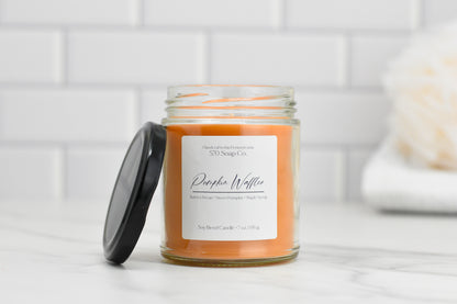 A glass jar labeled "Pumpkin Waffles Soy Blend Candle" by 570 Soap Co., filled with cozy aroma orange wax, rests on a white surface. The lid is placed beside it, with a background featuring white tile and a blurred object, possibly a towel or bath sponge.