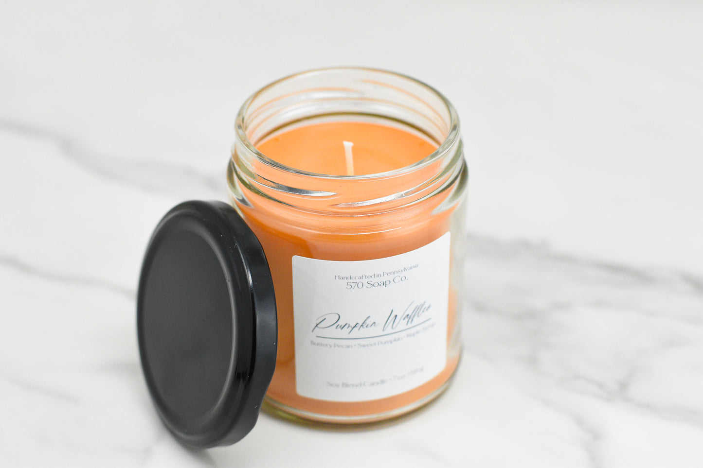 A jar candle with orange wax, named Pumpkin Waffles Soy Blend Candle from 570 Soap Co., fills the air with a cozy aroma. This soy blend candle, accompanied by a black lid, rests elegantly on a light marble surface.