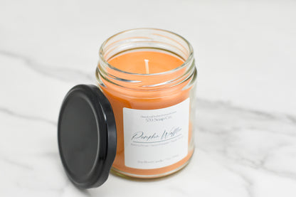 A jar candle with orange wax, named Pumpkin Waffles Soy Blend Candle from 570 Soap Co., fills the air with a cozy aroma. This soy blend candle, accompanied by a black lid, rests elegantly on a light marble surface.