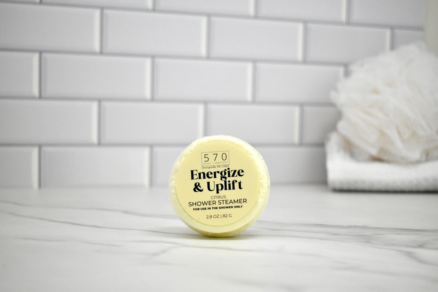 Shower steamer labeled 'Energize & Uplift' on a bathroom counter with tiled wall in the background.