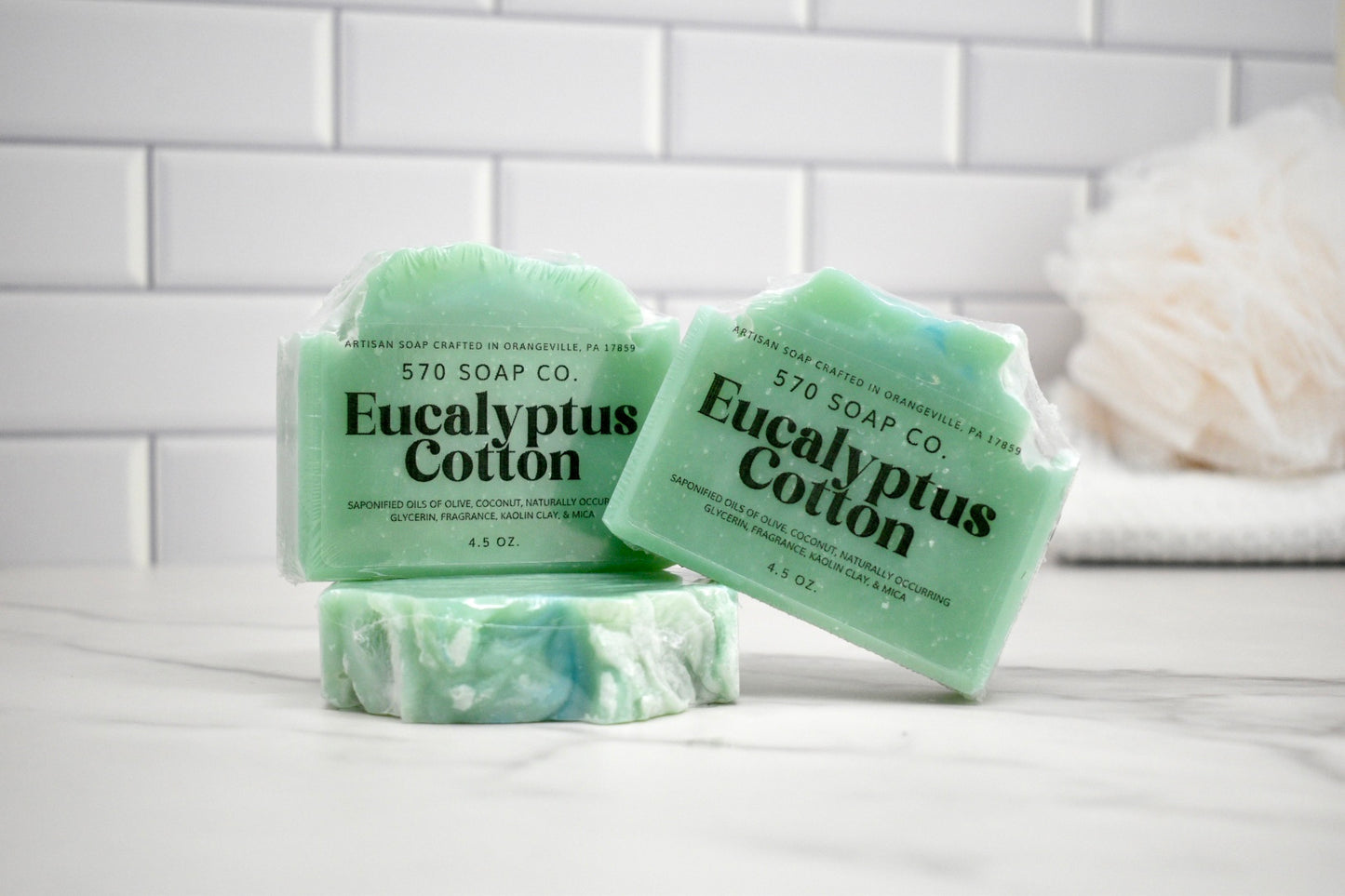 Three bars of Eucalyptus Cotton soap from 570 Soap Co on a white surface with a tiled wall background.