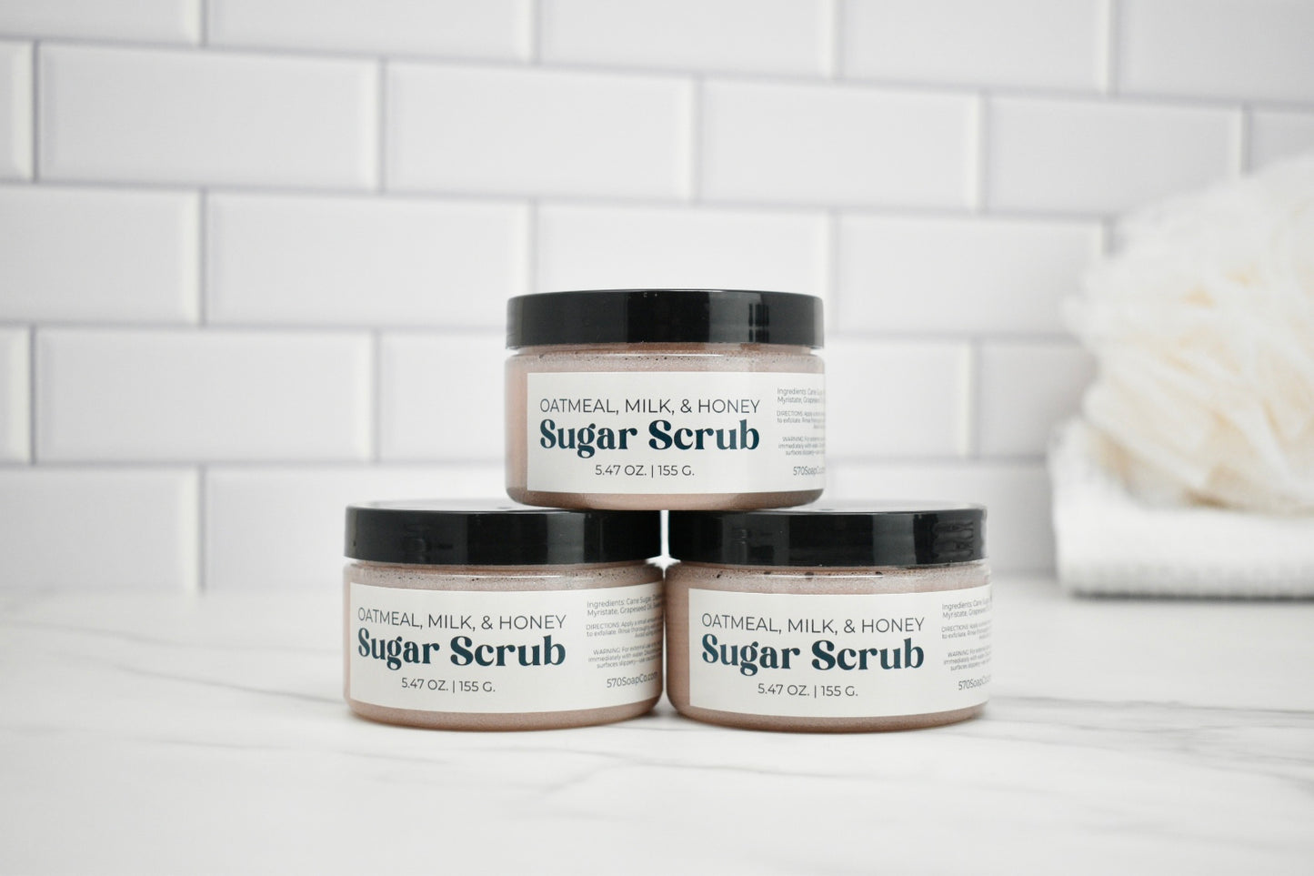 Three jars of Oatmeal Milk & Honey Sugar Scrub on a white surface with a tiled wall background.