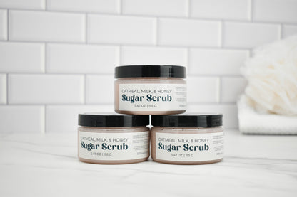 Three jars of Oatmeal Milk & Honey Sugar Scrub on a white surface with a tiled wall background.