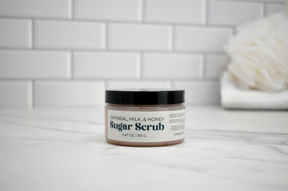 Jar of Oatmeal Milk & Honey Sugar Scrub on a bathroom counter with a tiled wall background.