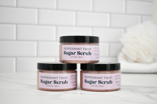 Three jars of peppermint twist sugar scrub stacked on a white surface with a tiled wall background.