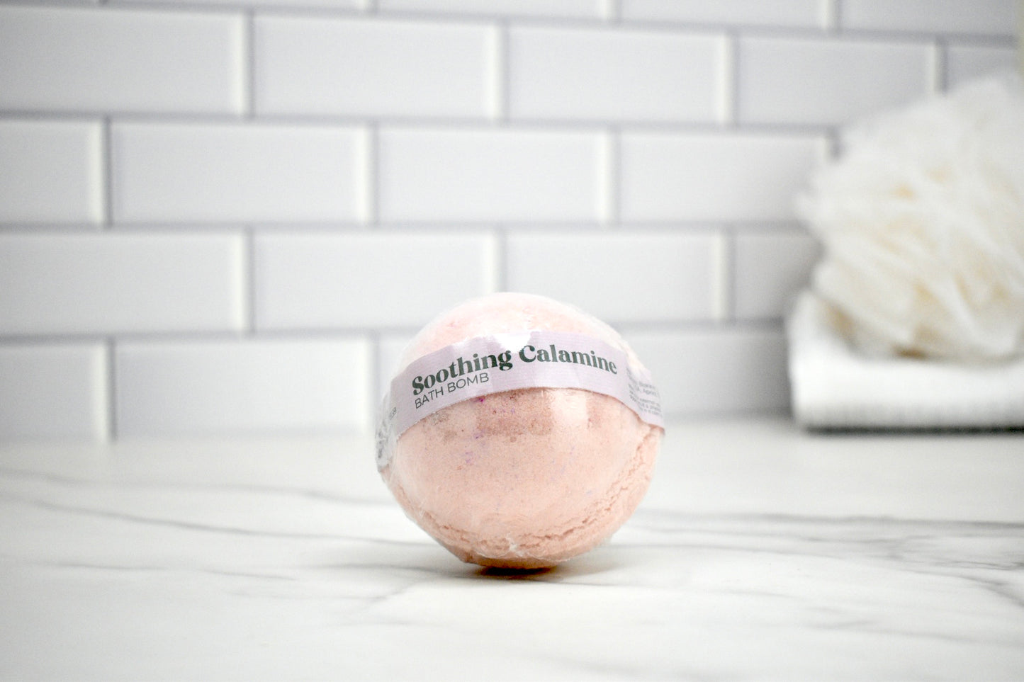 Bath bomb labeled 'Soothing Calamine' on a marble surface with a tiled wall background