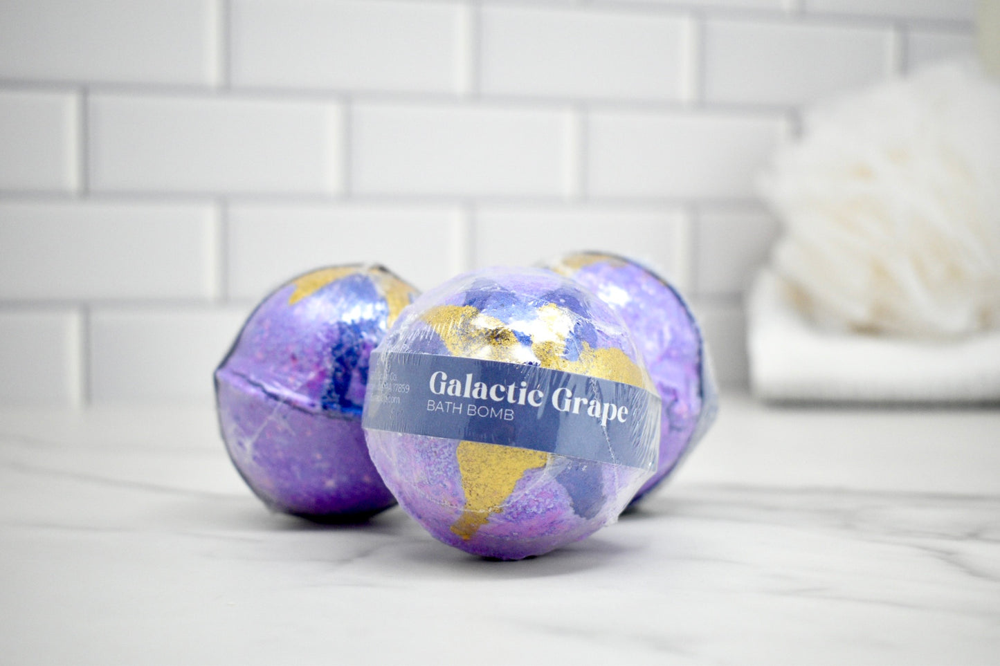 Two purple bath bombs labeled 'Galactic Grape' on a white surface with a tiled wall background.