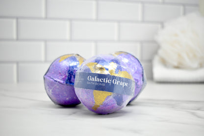 Two purple bath bombs labeled 'Galactic Grape' on a white surface with a tiled wall background.