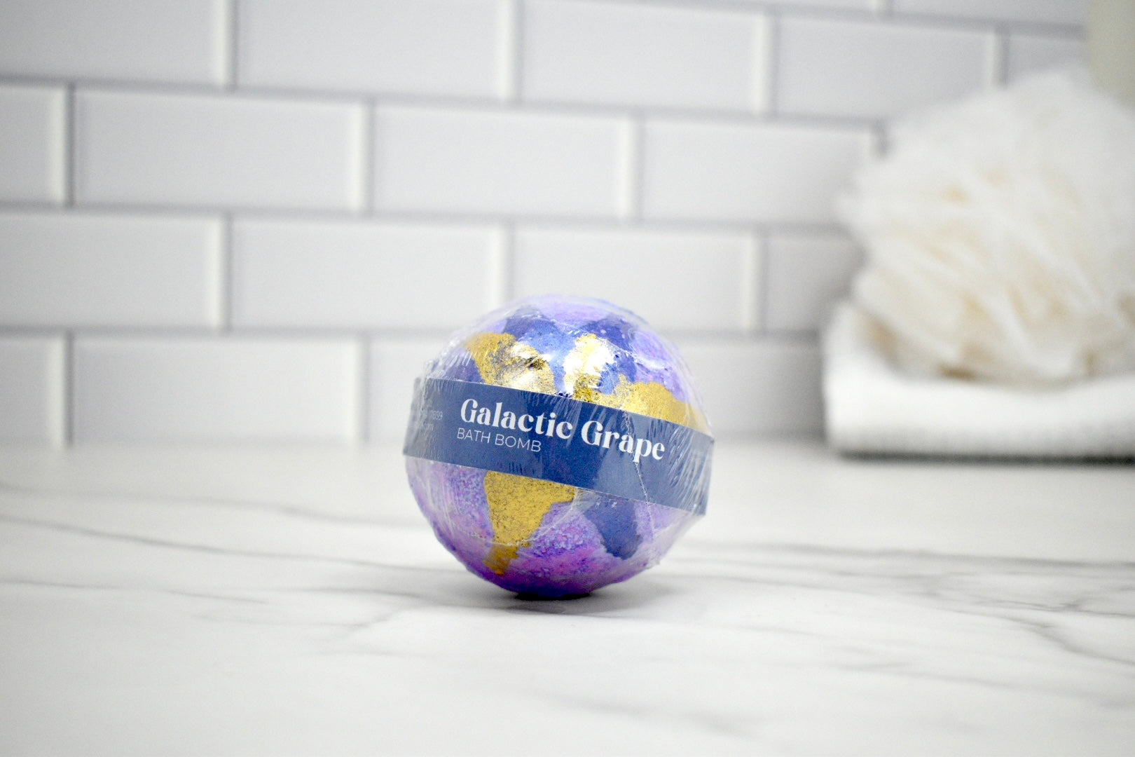 Bath bomb labeled 'Galactic Grape' on a tiled bathroom floor.