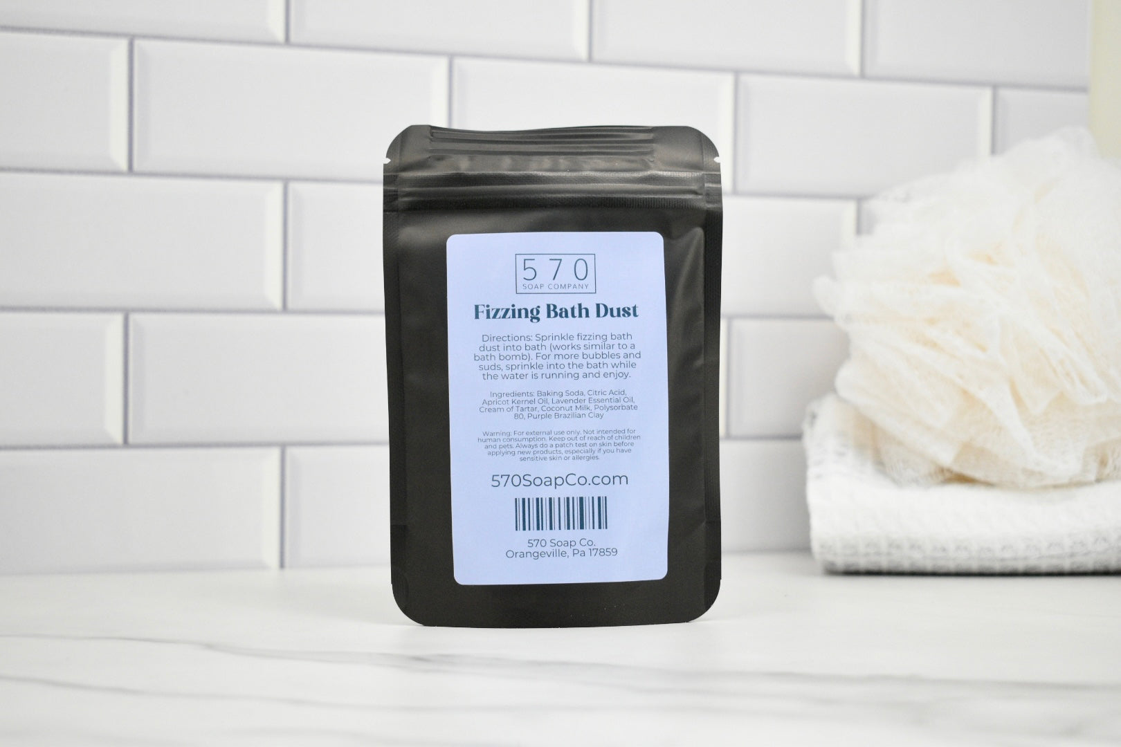 Black packaging of 570 Fizzing Bath Dust with a white label on a white tiled background.