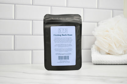 Black packaging of 570 Fizzing Bath Dust with a white label on a white tiled background.
