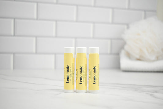 Three lip balm tubes labeled 'Lemonade' on a white surface with a tiled wall background.
