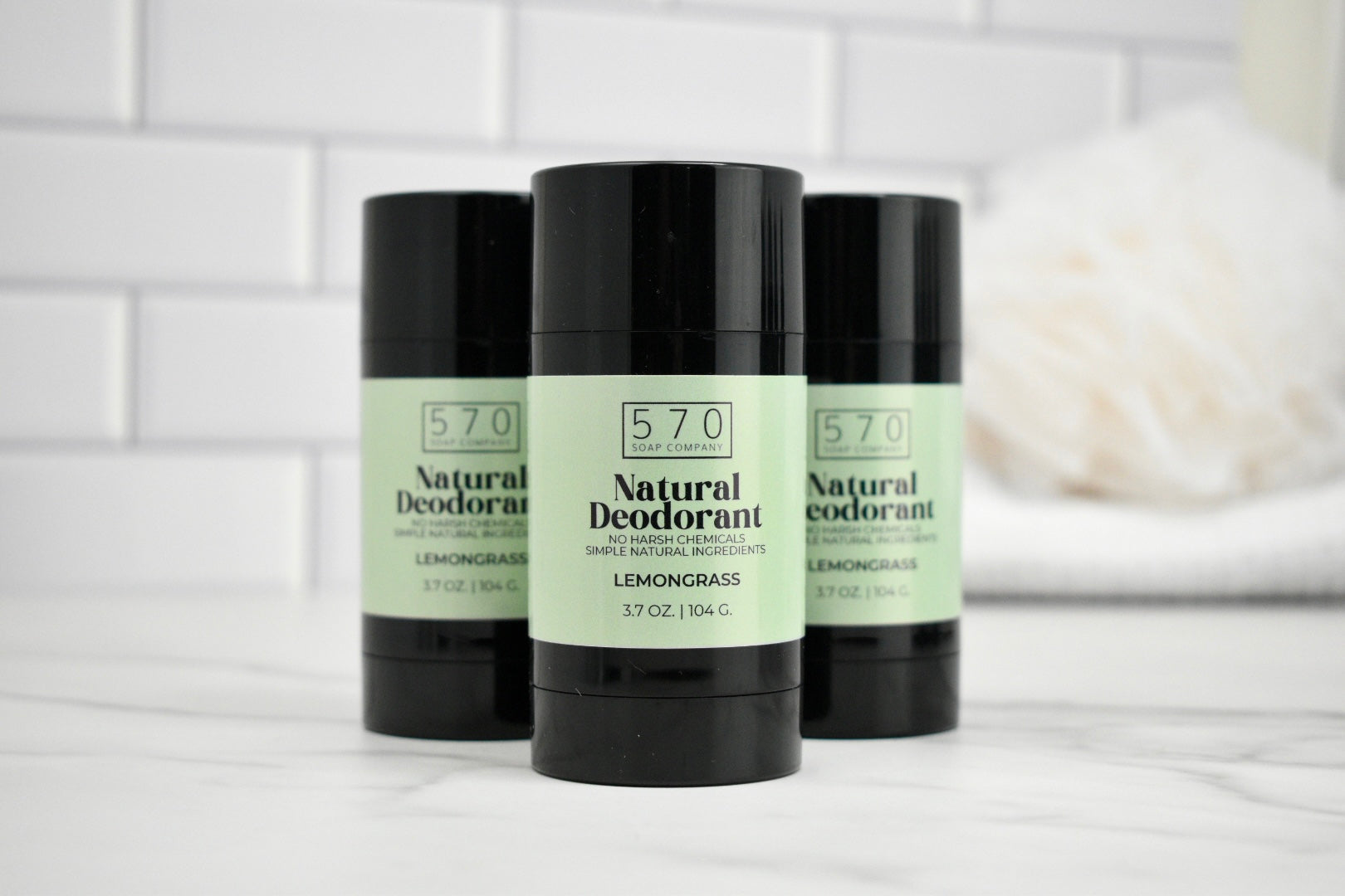 Three bottles of 570 Natural Deodorant on a white surface with a white tiled wall background.