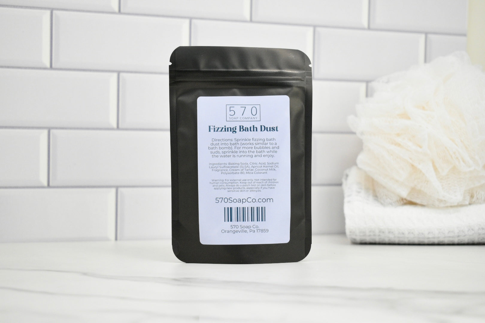 Black packaging of 570 Fizzing Bath Dust on a white surface with a tiled wall background.