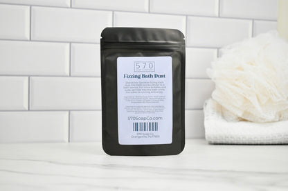 Black packaging of 570 Fizzing Bath Dust on a white surface with a tiled wall background.