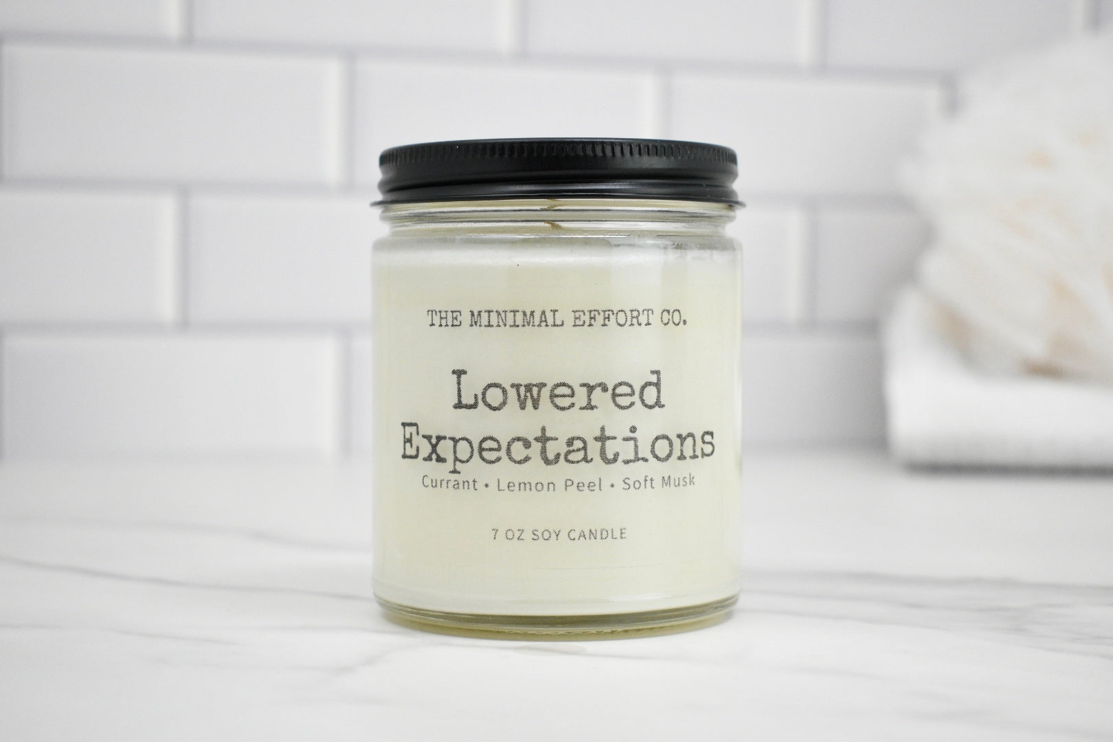 Candle jar labeled 'Lowered Expectations' by The Minimal Effort Co. on a white surface with a tiled wall background.