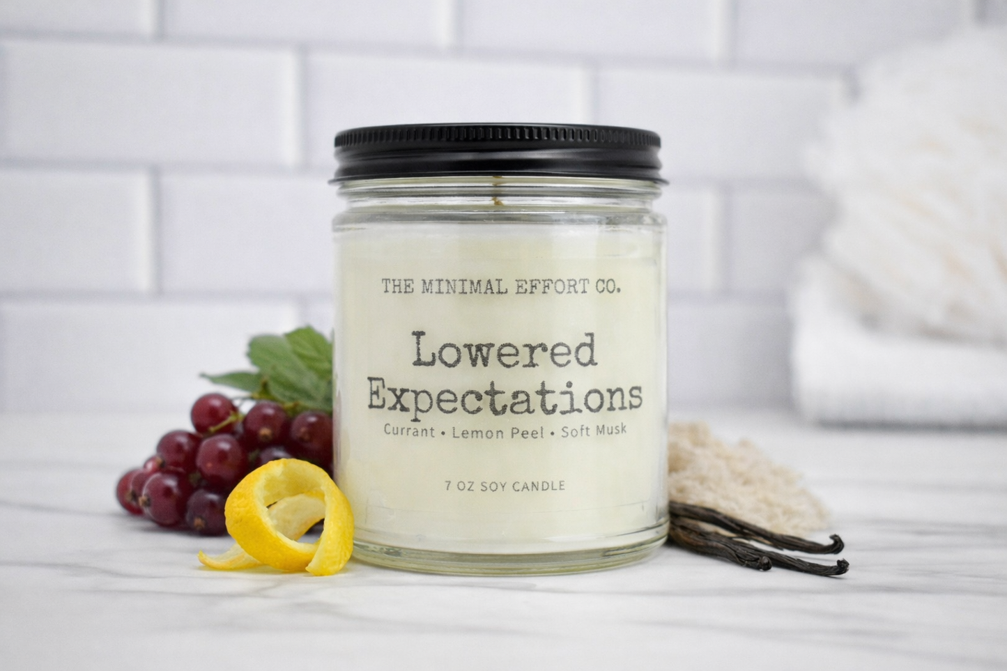 Candle jar labeled 'Lowered Expectations' from The Minimal Effort Co. on a white surface with grapes and vanilla beans.