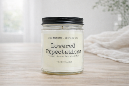 Candle jar labeled 'Lowered Expectations' by The Minimal Effort Co. on a neutral background.