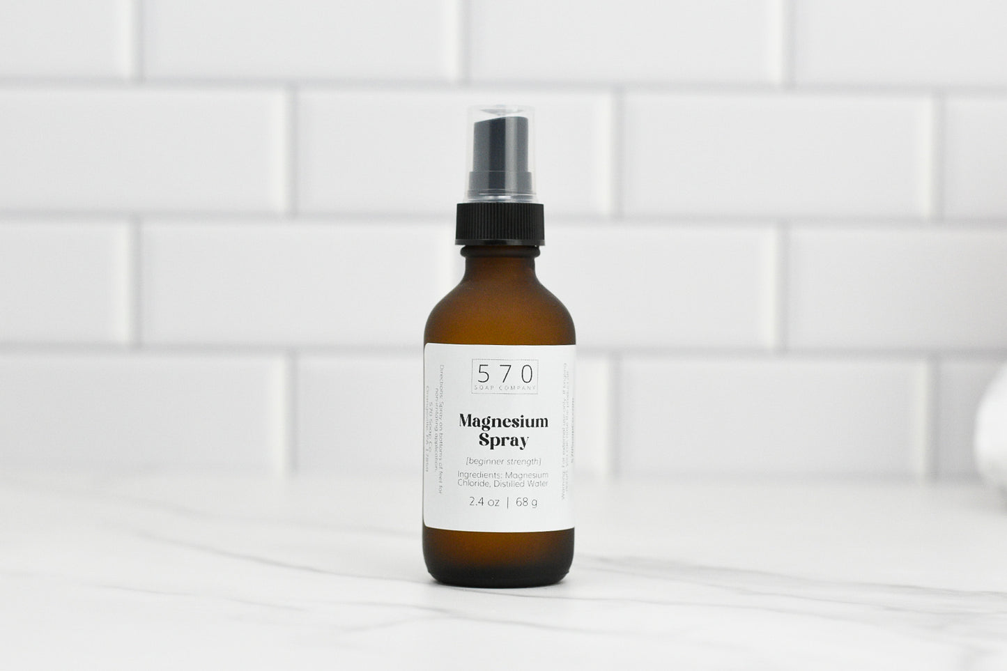 An elegant brown bottle of Magnesium Spray (beginner strength) from 570 Soap Co. features a sleek black pump top, gracefully positioned on a white countertop against a pristine tile backsplash. The label prominently displays the product's details.