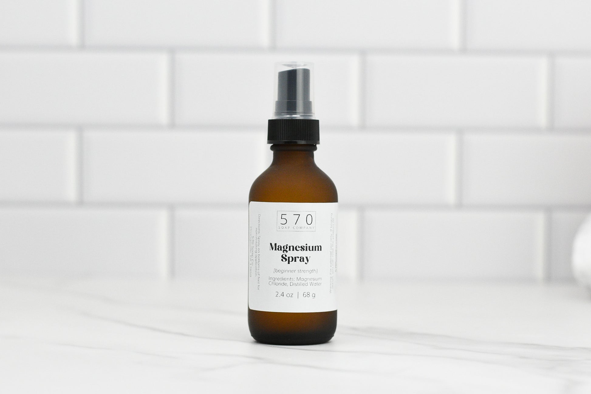 An elegant brown bottle of Magnesium Spray (beginner strength) from 570 Soap Co. features a sleek black pump top, gracefully positioned on a white countertop against a pristine tile backsplash. The label prominently displays the product's details.