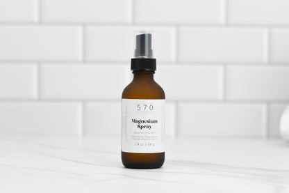 An elegant brown bottle of Magnesium Spray (beginner strength) from 570 Soap Co. features a sleek black pump top, gracefully positioned on a white countertop against a pristine tile backsplash. The label prominently displays the product's details.