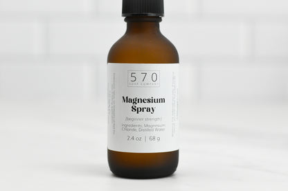 A brown bottle with a black cap labeled 570 Soap Co. Magnesium Spray (beginner strength). It contains magnesium chloride and distilled water, holding 2.4 oz or 68 g, placed on a white surface.