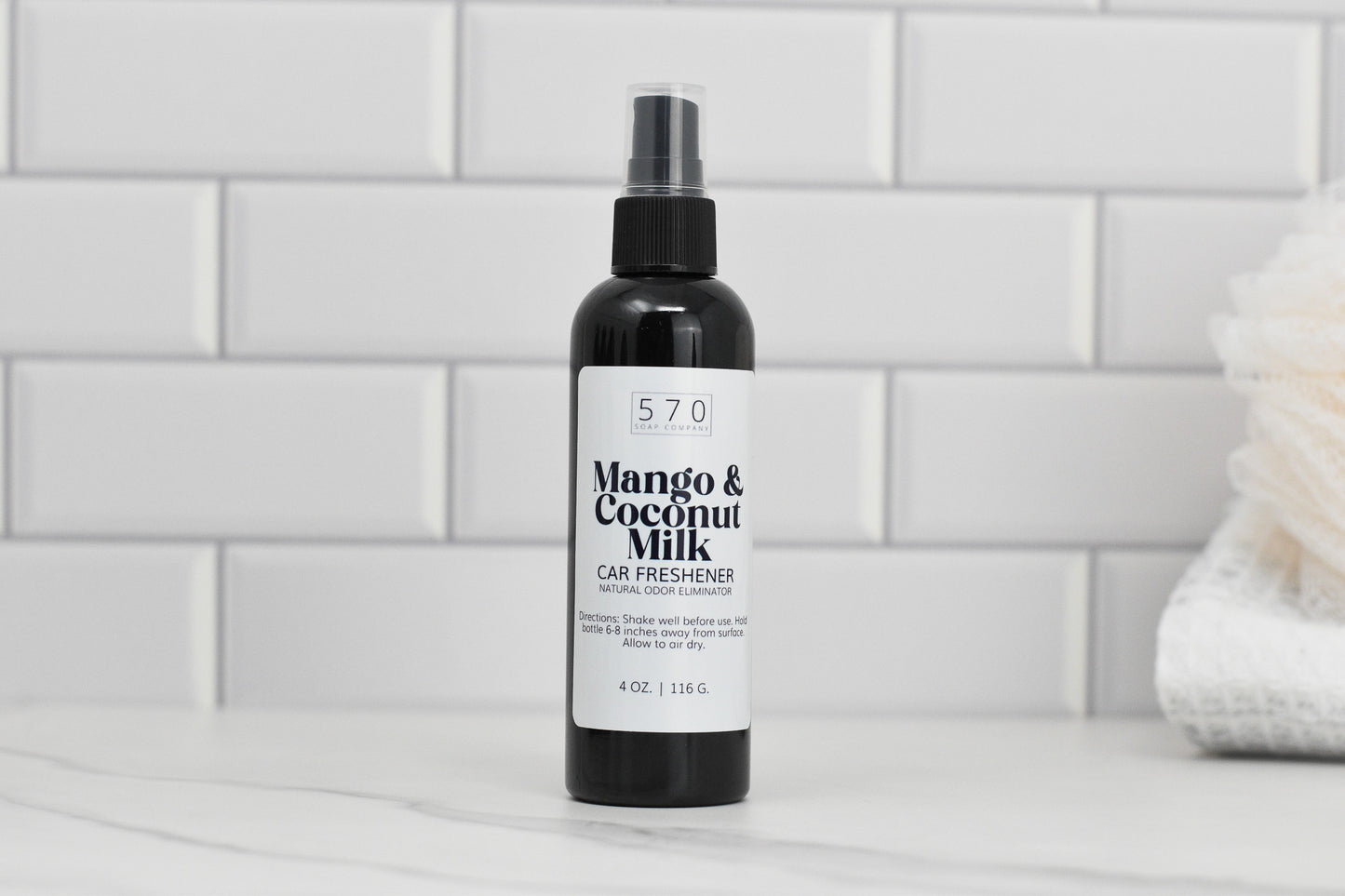 A black bottle labeled Linen & Room Spray 4 oz from 570 Soap Co., known for its natural odor eliminating properties, sits on a white marble countertop. The background features white subway tiles and a soft, folded towel on the right.