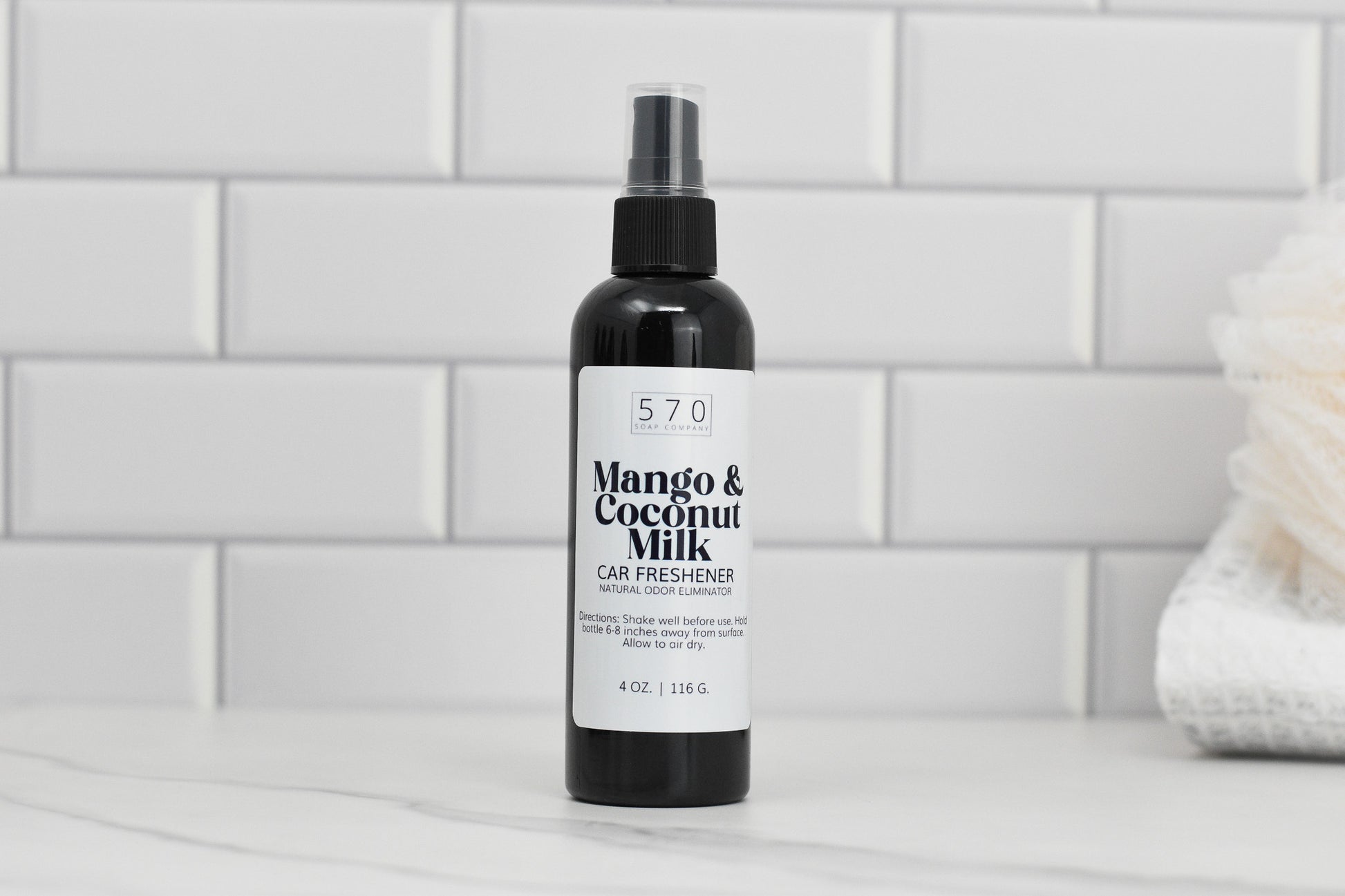 A black bottle labeled Linen & Room Spray 4 oz from 570 Soap Co., known for its natural odor eliminating properties, sits on a white marble countertop. The background features white subway tiles and a soft, folded towel on the right.