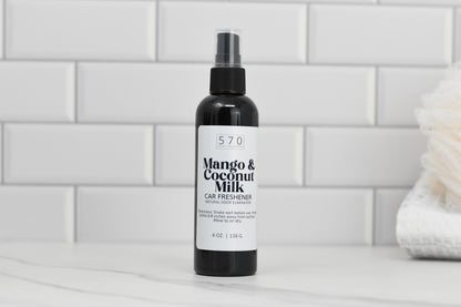 A black bottle labeled Linen & Room Spray 4 oz from 570 Soap Co., known for its natural odor eliminating properties, sits on a white marble countertop. The background features white subway tiles and a soft, folded towel on the right.