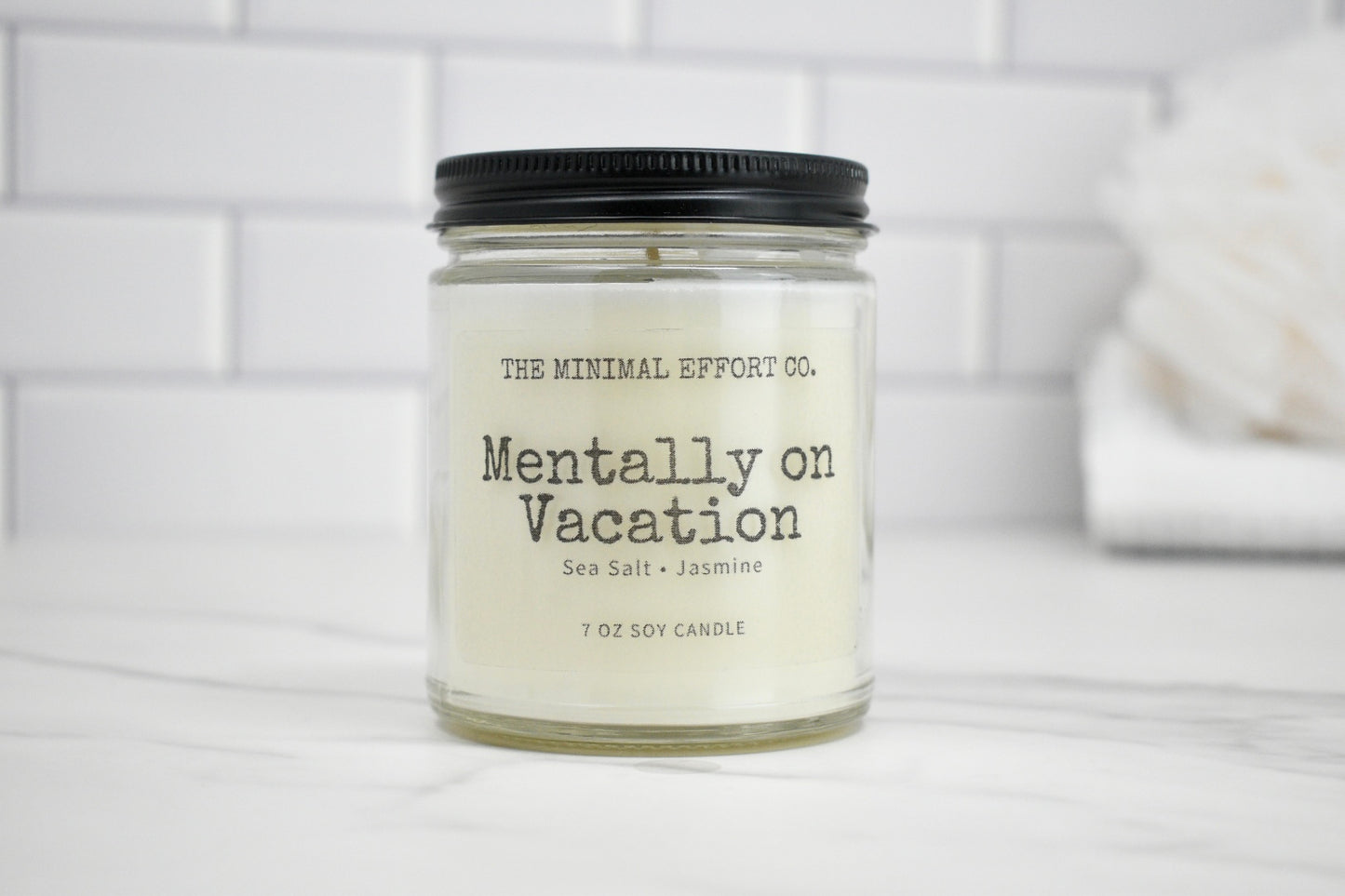 Candle labeled 'Mentally on Vacation' by The Minimal Effort Co. on a white surface with a tiled wall background.