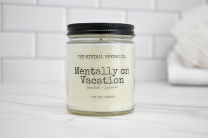 Candle labeled 'Mentally on Vacation' by The Minimal Effort Co. on a white surface with a tiled wall background.