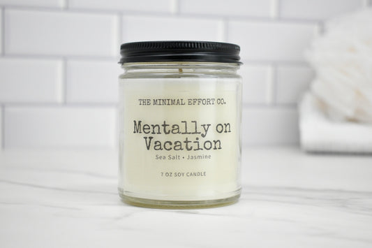 Candle labeled 'Mentally on Vacation' by The Minimal Effort Co. on a white surface with a tiled wall background.