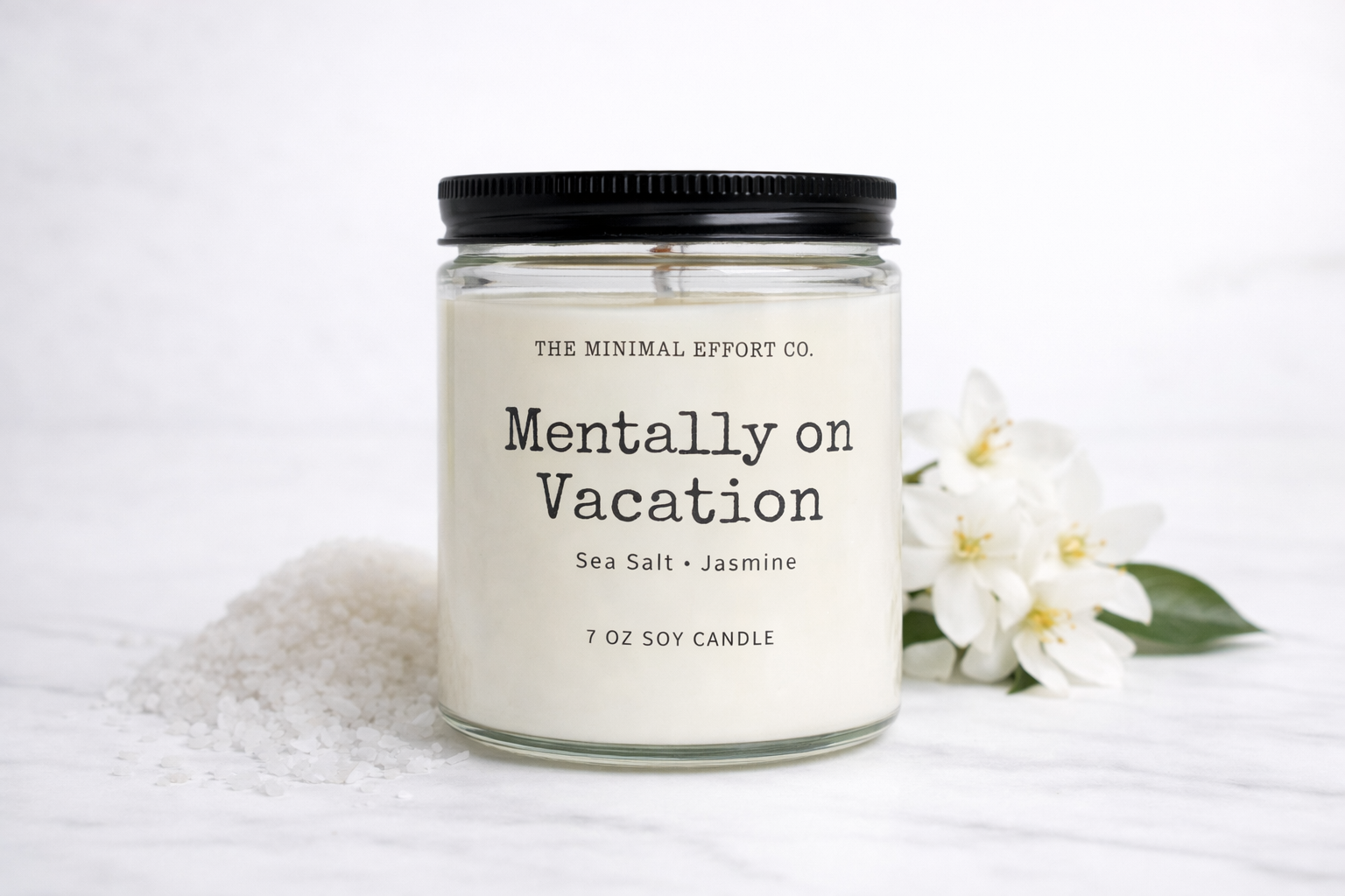 Candle labeled 'Mentally on Vacation' from The Minimal Effort Co. with a white flower and towel in the background.