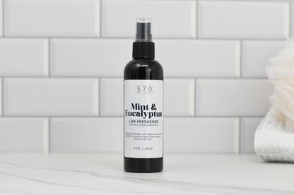 A black bottle labeled 570 Soap Co. Linen & Room Spray 4 oz Plastic Bottle rests on a countertop against a white subway tile backdrop, emitting refreshing fragrances. A cream-colored cloth is partially visible on the right, suggesting its dual use for natural odor elimination as both a Linen & Room Spray.