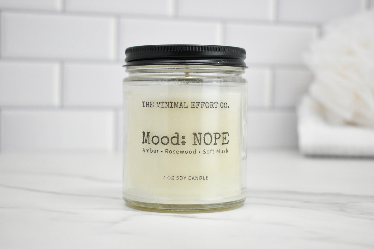 Candle jar labeled 'Mood: NOPE' by The Minimal Effort Co. on a white surface with a tiled wall background.