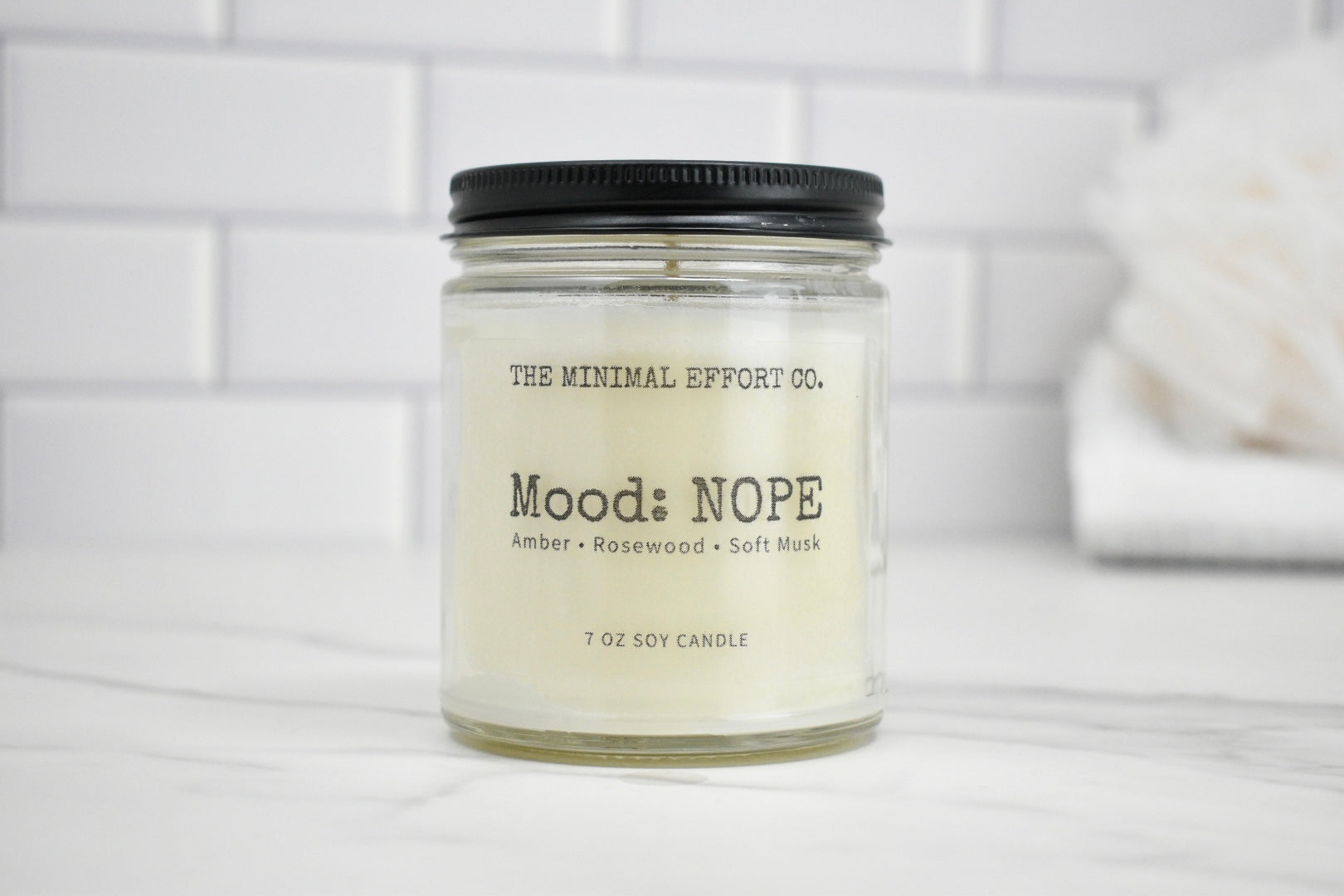Candle jar labeled 'Mood: NOPE' by The Minimal Effort Co. on a white surface with a tiled wall background.